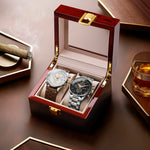 Two watches in a wooden box with a glass lid, surrounded by a glass of whiskey and a cigar on a wooden surface.