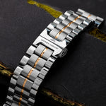 A silver watch band with 18K gold-plated details, featuring a clasp and links, placed on a textured dark surface.