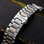 A silver watch band with 18K gold-plated details, featuring a clasp and links, placed on a textured dark surface.
