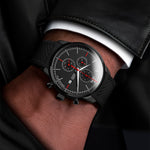 Black wristwatch with a detailed red face on a person's wrist, wearing a black leather jacket.