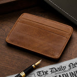 Brown leather cardholder on a wooden surface with a pen and newspaper.